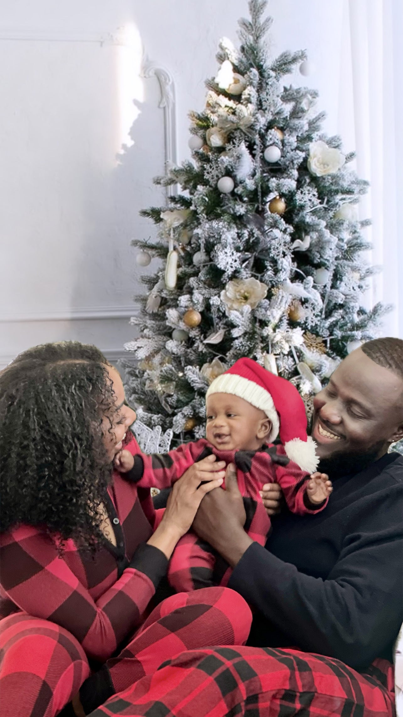 Family of three in matching red and black pajamas sitting in front of a decorated Christmas tree.